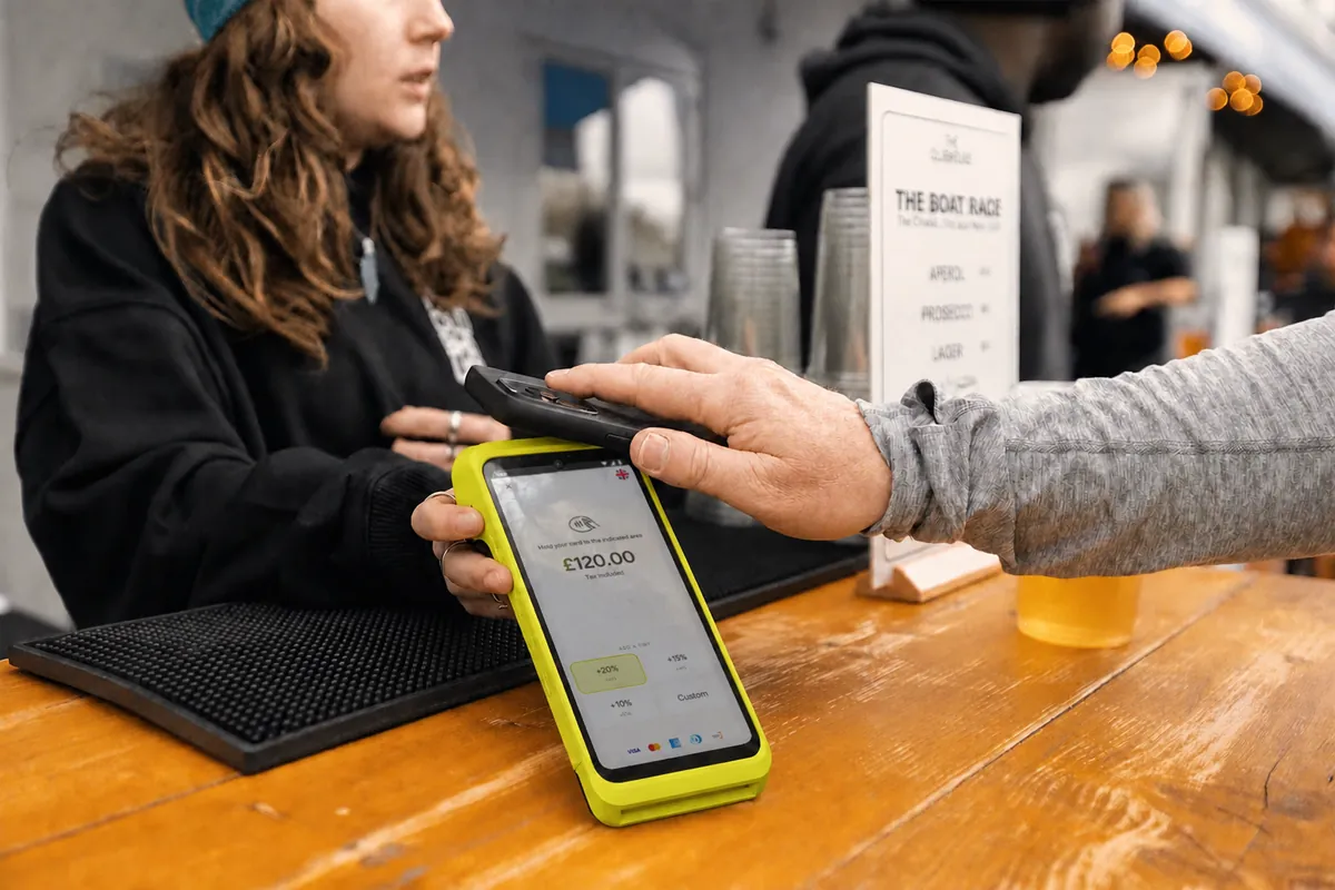 A barista hands a Teya Pro across a wooden cafe counter while the customer reaches to tap a card.